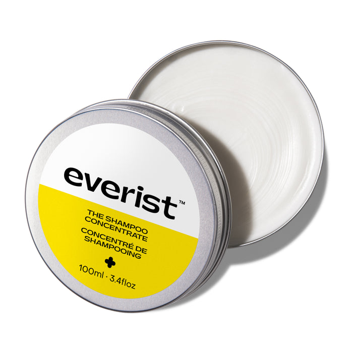 A round metal tin with a partially opened lid reveals creamy white shampoo inside. The label reads Everist The Shampoo Concentrate Tin, featuring bond repair technology, net weight 100ml / 3.4 fl oz.