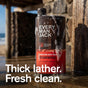 A bottle of Every Man Jack Hydrating Body Wash for Men stands upright, covered in suds with water pouring over it. The beach background features a wooden structure. Text reads: Thick lather. Fresh clean. Made with naturally-derived ingredients.
