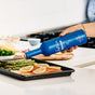 A hand pours Kosterina Organic Everyday Extra Virgin Olive Oil from a blue bottle over salmon, asparagus, and lemon slices on a baking tray in a modern kitchen.