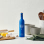 A blue bottle of Kosterina Organic Everyday Extra Virgin Olive Oil sits on a kitchen counter beside mixing bowls, a green towel, cracked eggs in a bowl, eggshells, a rolling pin, and utensils against a white background.