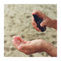 A person sprays Everyday Oil, Mainstay Blend by Everyday Oil from a small brown bottle onto their open palm, with a blurred sandy background visible.