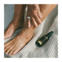 While seated on a textured, light-colored fabric, a person applies Everyday Oil’s Early Morning Blend to their lower leg and ankle. The dark glass bottle sits nearby, offering botanical oils and microbiome-friendly skincare benefits.