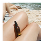 A bottle of Everyday Oil, Mainstay Blend by Everyday Oil rests on sunlit legs by the water, while another person stands near rocky shoreline in the background.