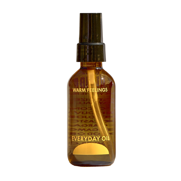 A brown glass bottle with a black spray cap labeled Everyday Oil, Warm Feelings Blend by Everyday Oil in gold text, featuring botanical oils for microbiome-friendly skincare, is photographed on a white background.