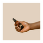 A hand holds a small brown bottle with a black pump dispenser labeled Everyday Oil, Warm Feelings Blend by Everyday Oil, shown against a beige background. The bottle contains an organic skincare oil infused with botanical ingredients.