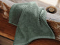 A green Coyuchi Air Weight® Organic Towel with a textured finish is draped over the edge of a stone sink, revealing a wooden surface below.