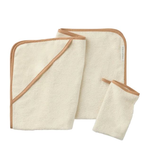 Three cream-colored bath items with tan edging from Coyuchi’s Cloud Loom™ Organic Baby Hooded Towel and Bath Mitt Set—including a hooded towel, washcloth, and bath mitt—are laid flat on a white surface.