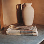 A ceramic jug sits on a stone counter next to folded Coyuchi Miraloma Organic Kitchen Towels (set of 4), three clear glasses, and a wooden cutting board, all in warm, soft lighting.