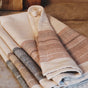 A folded stack of the Coyuchi Miraloma Organic Kitchen Towels, Set of 4, in beige and striped designs, rests on a brown countertop with a wooden cutting board and glass in the background.