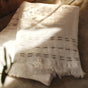 A set of two Alder Organic Hand Towels by Coyuchi, crafted from artisanal off-white cotton with fringe edges and stitched patterns, is displayed on a light surface, its gauze texture accentuated by soft sunlight and gentle shadows.