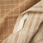 Close-up of beige fabric with white grid and stripe patterns, featuring a folded edge and a partially visible off-white tag; crafted from GOTS certified cotton, the Clarendon Organic Kitchen Towels by Coyuchi combine elegance with eco-conscious quality in a sustainable set of four.
