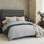 A modern bedroom featuring a gray upholstered bed, Coyuchi’s Organic Crinkled Percale™ Duvet Cover in soothing gray tones, neutral pillows, a wooden nightstand with a lamp, and green plants placed by a textured wall and large window.