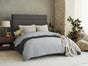 A modern bedroom featuring a gray upholstered bed dressed in layered bedding, including Coyuchi’s Organic Crinkled Percale™ Sham and an Organic Crinkled Percale Sheet, complemented by a washed cotton sham. A bedside table with a lamp, indoor plants, and neutral decor sits against a textured stone and plaster wall. All textiles are crafted from GOTS Certified organic cotton for optimal comfort and sustainability.