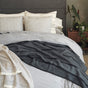 A neatly made bed is dressed in gray GOTS Certified organic cotton bedding with pillows, next to a wooden nightstand topped with potted plants. More greenery and a stone accent grace the right side of the room, while the Coyuchi Organic Crinkled Percale™ Duvet Cover lends breathable comfort to this inviting space.
