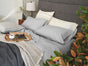 A cozy bed is layered with gray sheets and topped with Coyuchi’s Organic Crinkled Percale™ Pillowcases. A wooden tray rests on the bed, holding a coffee cup, saucer, glasses, and a notebook. In the foreground sits a plant, while a lamp and pillow are arranged on the bedside table.