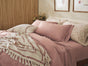 A neatly made bed features Coyuchi’s Organic Crinkled Percale™ Sheet Set in pink, paired with patterned pillows and a fringed throw blanket. Framed wall art hangs above the bed, while dried flowers rest on a bedside table, creating a cozy and inviting atmosphere enhanced by the breathable Coyuchi bedding.