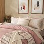 A neatly made bed, dressed in pink and floral bedding, features a striped pillow and a textured throw as highlights. Taking center stage is the Coyuchi Organic Crinkled Percale™ Duvet Cover, adding comfort with its organic cotton fabric. Two abstract art pieces hang above the bed, while a vase of dried flowers adorns the nightstand.