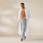 A man stands barefoot on a light-colored floor, smiling and looking down, wearing the Coyuchi Unisex Organic Waffle Robe in white over gray pajama pants against a plain beige background.