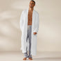 A man stands smiling, barefoot, against a plain light-colored background, wearing the Coyuchi Unisex Organic Waffle Robe in white over gray pajama pants.