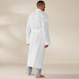 A man stands barefoot on a plain floor, facing away. He is wearing the Coyuchi Unisex Organic Waffle Robe with a belt over light gray pants. The background is neutral and uncluttered.