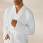A person is shown from the shoulders to mid-thigh wearing the Coyuchi Unisex Organic Waffle Robe, with one hand adjusting the robe and the other resting in a pocket against a plain, neutral background.