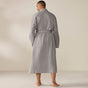 A man is seen from behind, barefoot, wearing the Coyuchi Unisex Organic Waffle Robe in gray with a belted waist, against a neutral beige background.