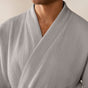 A person wearing the Coyuchi Unisex Organic Waffle Robe in light gray, featuring a textured waffle weave, is shown from the shoulders to upper chest with their face mostly out of frame against a plain background.