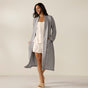 A woman smiles, wearing the Coyuchi Unisex Organic Waffle Robe in gray over a white floral cami and shorts set, paired with fluffy tan slippers.