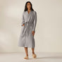 A woman stands against a neutral background wearing the Coyuchi Unisex Organic Waffle Robe in gray, featuring a long, classic silhouette with a waffle-weave texture, belt closure, and front pockets. She pairs it with fluffy tan slippers for an effortlessly cozy and stylish lounge look.