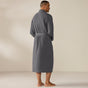 A man stands barefoot, facing away, wearing the Coyuchi Unisex Organic Waffle Robe in gray, belted at the waist, in a neutral-colored room with a plain background.
