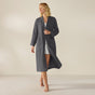 A woman stands barefoot in the Coyuchi Unisex Organic Waffle Robe in grey, layered over light sleepwear. She smiles and gazes to the side with one leg slightly forward, set against a plain beige background.