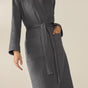 A person wears the Coyuchi Unisex Organic Waffle Robe—a charcoal grey, long-sleeve robe made from soft organic cotton with a tie waist and front pockets—standing out against a plain, light beige background.