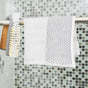 A Full Circle Tidy Dish Cloth, made from white organic cotton with a black dotted pattern and non-toxic dyes, hangs on a metal bar beside a dish brush with white bristles and a bamboo handle against a mosaic-tiled wall.