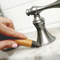 A hand with light pink nail polish uses the Full Circle Micro Manager Detail Brush with a wooden handle to scrub around the base of a silver bathroom faucet on a marble sink.