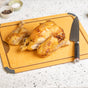 A roasted chicken, partially carved with one leg and one wing removed, sits on a cutting board next to a chef’s knife. Small bowls of salt and pepper are in the background on a speckled countertop.