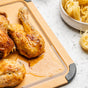 Roast chicken pieces with golden, crispy skin rest on a cutting board, juices pooling around them. In the background, uncooked fettuccine pasta nests sit in a white bowl on a light countertop.