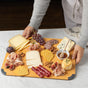 A person holds a rectangular wooden board with assorted cheeses, crackers, cured meats, figs, grapes, nuts, and a small bowl of honey with a dipper, arranged neatly on a marble surface.