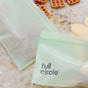 Two green Full Circle Compostable Zipper Snack Bags, one with pretzel pieces and the other with apple slices, sit on a marble countertop. The front bag displays the Full Circle brand name.