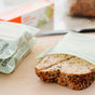 Two slices of multigrain bread sit partially inside a Full Circle Compostable Zipper Sandwich Bag on a kitchen counter, with a blurred bread loaf and box in the background.