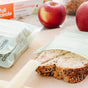 Two apples, a loaf of multigrain bread, a cheese stick, and a sandwich in a Full Circle Compostable Zipper Sandwich Bag rest on the counter. In the background, the box of these eco-friendly bags completes the scene.