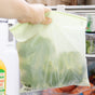 A hand holds a Full Circle Compostable Zipper Gallon Bag filled with leafy vegetables in front of an open fridge with orange juice and other produce visible inside.