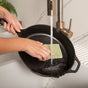 A person uses a Full Circle Agave Leaf Scrubber Sponge to clean a black cast iron skillet under running water, demonstrating proper cast iron care.