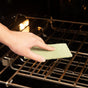 A hand is scrubbing an oven rack with a Full Circle Agave Leaf Scour Pad, cleaning the metal bars under warm light.