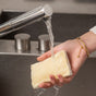 A hand holds a Full Circle 2pk Loofah Sponge under running water at a kitchen sink, droplets splashing onto the plastic-free, plant-based scrubber. The person wears a gold bracelet.