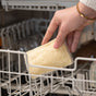 A hand places a 2pk Loofah Sponge by Full Circle, made from natural loofah and plastic-free, on the rack of an open dishwasher. The person wears a white sweater and gold bracelet; dishwasher interior and dishes are visible.