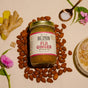 A jar of Big Spoon Roasters Fiji Ginger Almond Butter sits on a beige background, surrounded by whole almonds, fresh ginger, crystallized ginger, salt, and pink flowers.