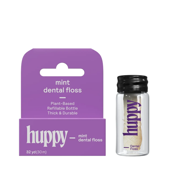A purple Huppy Vegan Mint Floss box stands next to a clear glass bottle with a black cap containing peppermint-infused, compostable floss. The bottle’s label matches the box design.