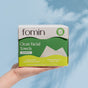 A hand holds a box of Fomin Clean Facial Towels Bamboo for sensitive skin, showcasing labels on anti-microbial and acne-fighting benefits. The background is soft blue with a subtle shadow.
