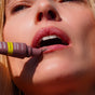 A close-up shows someone applying FRÉ Skincare’s PLUMP ME Peptide Lip Tint Mineral SPF 30 to their lips, the tube near their slightly open mouth, with smooth skin and natural complexion highlighted by the lighting.