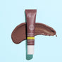 A tube of FRÉ Skincare PLUMP ME Peptide Lip Tint Mineral SPF 30 rests on a light blue background atop a smear of the brownish, lip-plumping formula.